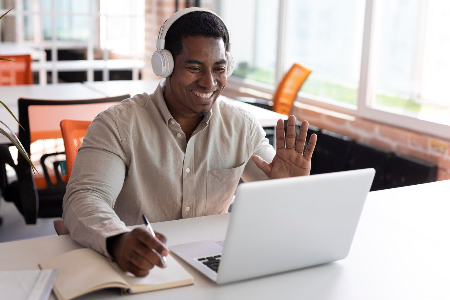 A man wearing headphones seated at a desk with a laptop open and waving to the video call recipient.